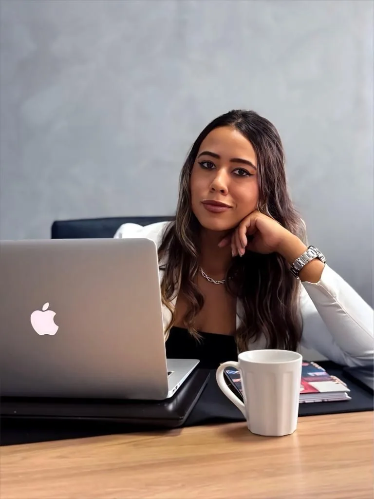Amanda Terra à mesa, com o notebook aberto e uma xícara ao lado, apoiando o rosto na mão em um momento de reflexão